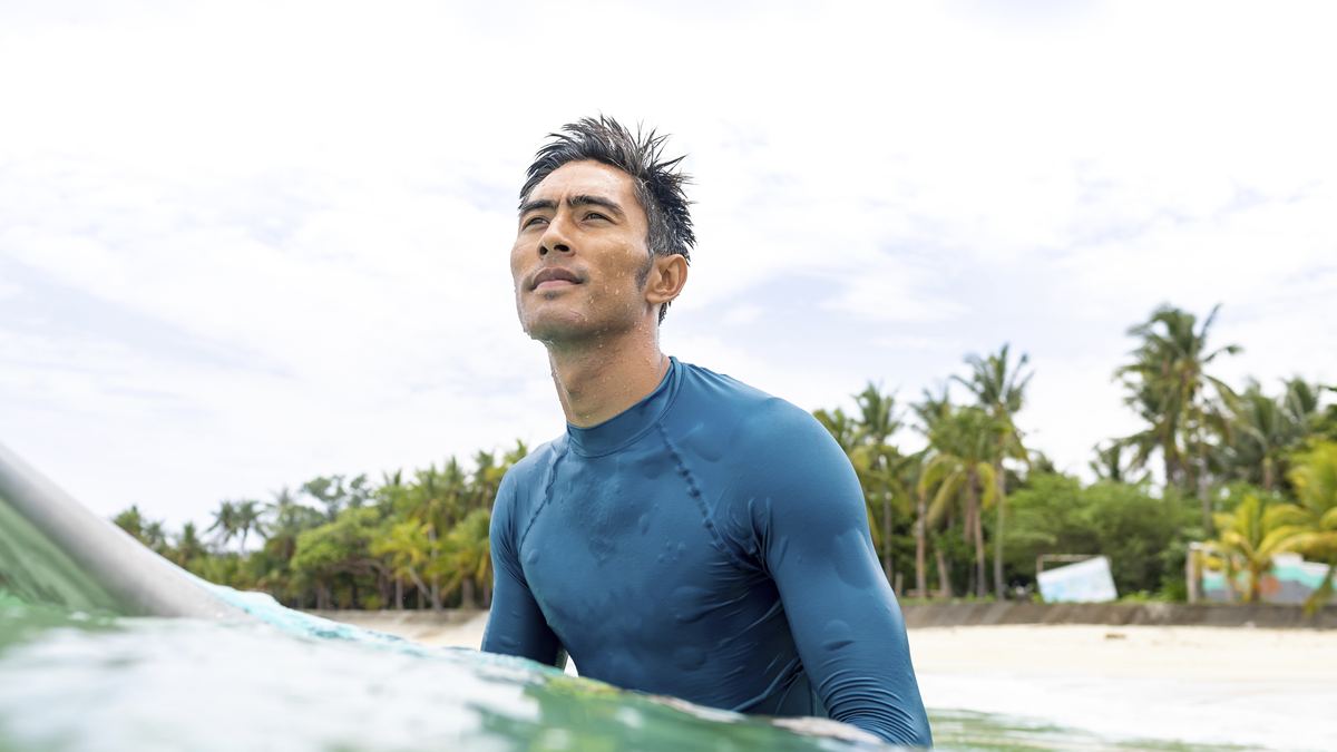 A man wearing a long-sleeve rash guard floating in the ocean near a tropical beach with palm trees, full sun coverage with no sunscreen needed on covered skin